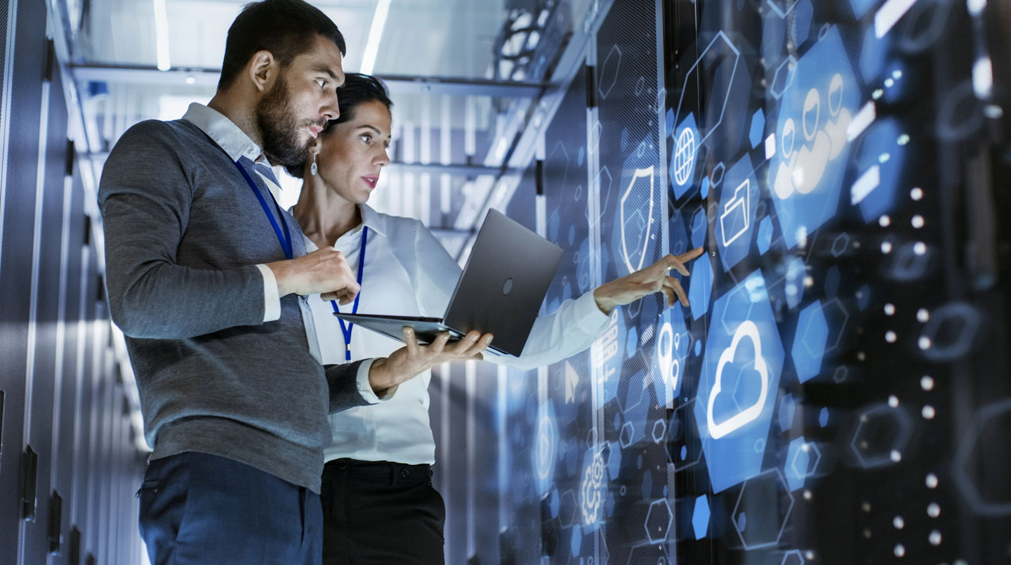 Technicien informatique de dos devant un mur de serveurs en datacenter, tenant un laptop et analysant l'infrastructure.